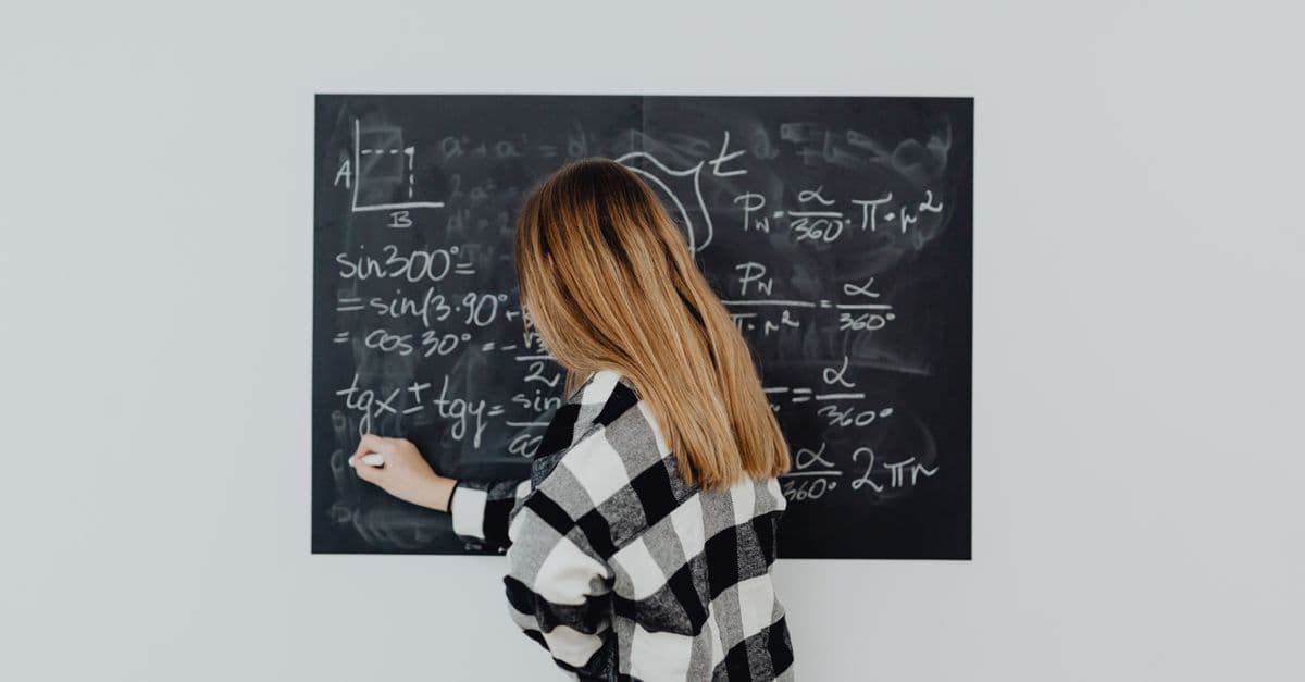 Teen girl in plaid shirt solving math equations on a blackboard, focusing on trigonometry.
