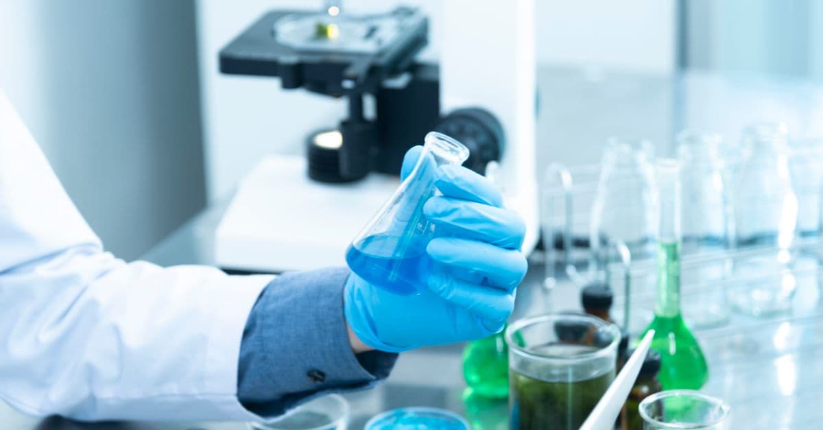 Scientist in gloves analyzing blue liquid in a laboratory setting with microscope and glassware.