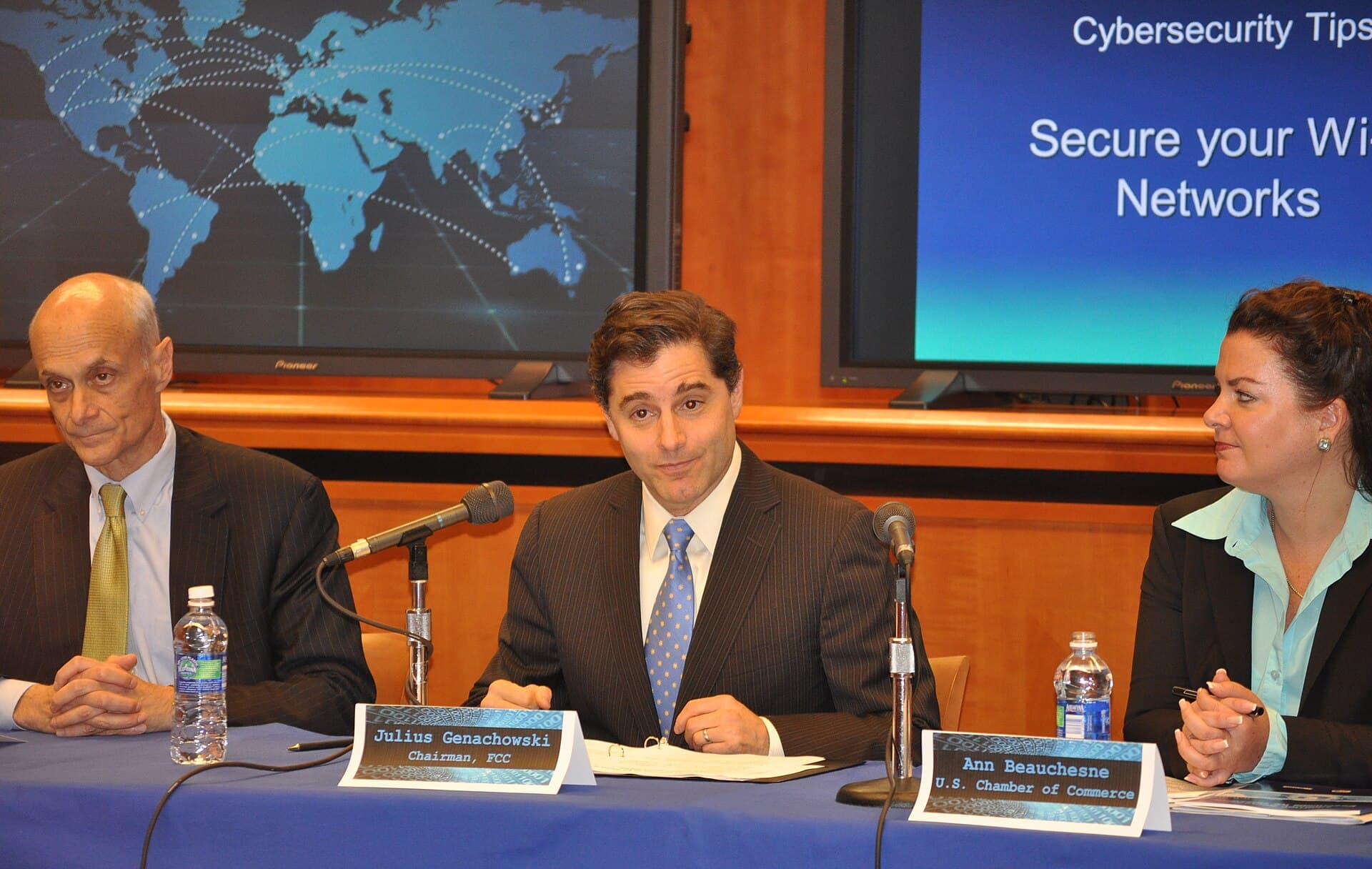 A public cybersecurity roundtable with speakers and a security-monitoring display in the background.