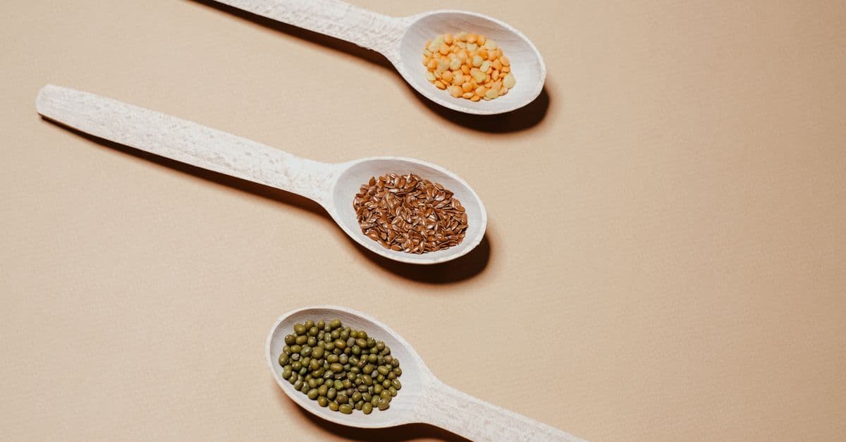 Flat lay of mung beans, flaxseeds, and split peas in wooden spoons, showcasing natural grains.