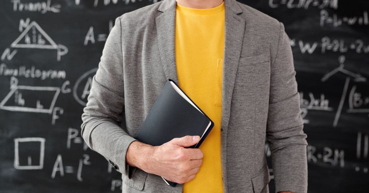 Close-up of a teacher holding a notebook in a classroom setting with equations on the blackboard.