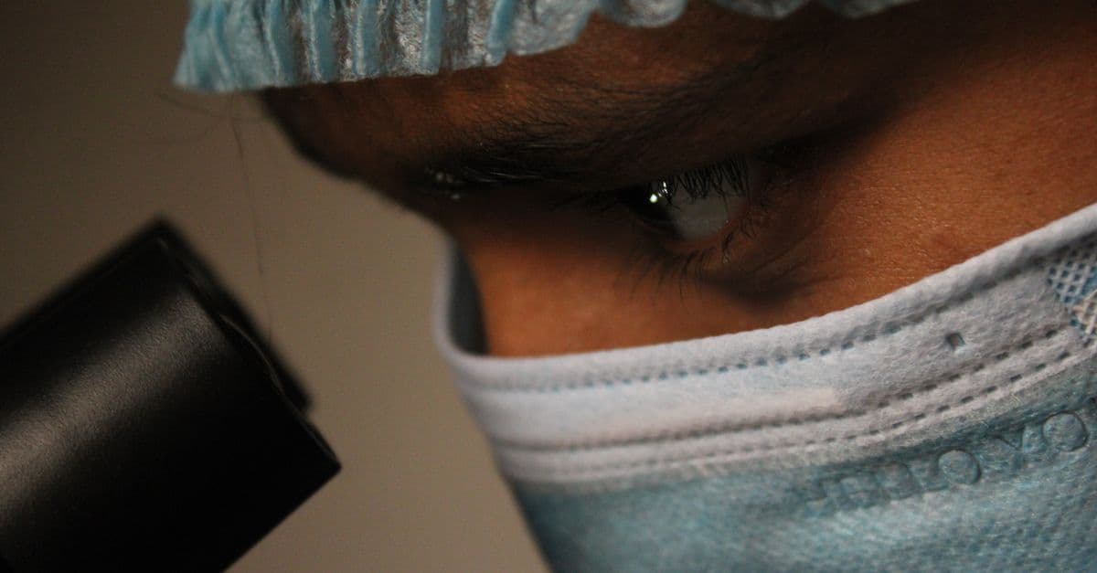 Close-up of a scientist wearing a mask and cap observing through a microscope.