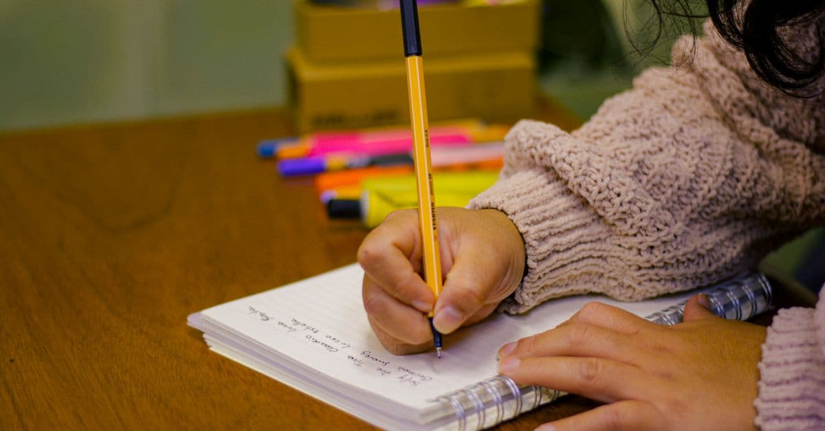 A person writing in a notebook with pens and highlighters on a wooden desk, creating a study scene.