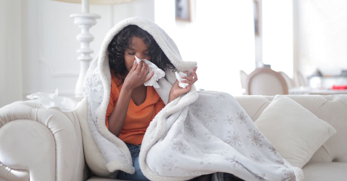 Sick young ethnic lady in casual clothes sitting on comfortable sofa under warm blanket and blowing nose into paper handkerchief at home