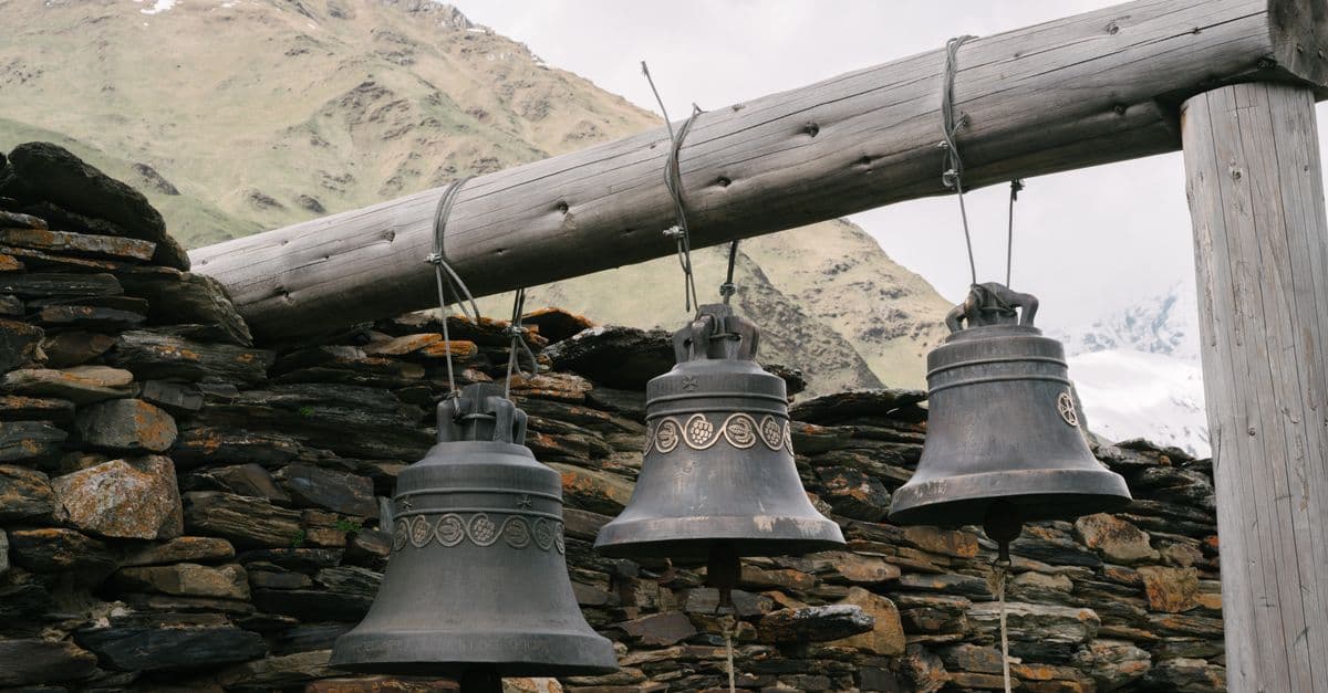 Three ornate bells hang on a stone wall with a mountain backdrop, showcasing rustic charm.