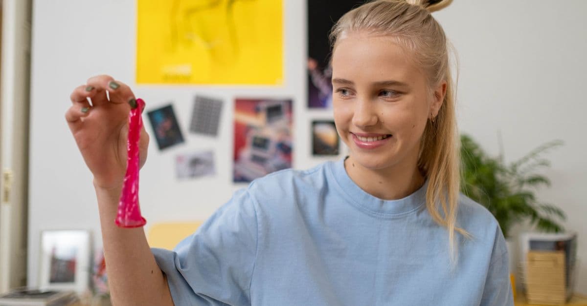 A smiling teenager in a bedroom holding a pink contraceptive device, expressing safe sex education.