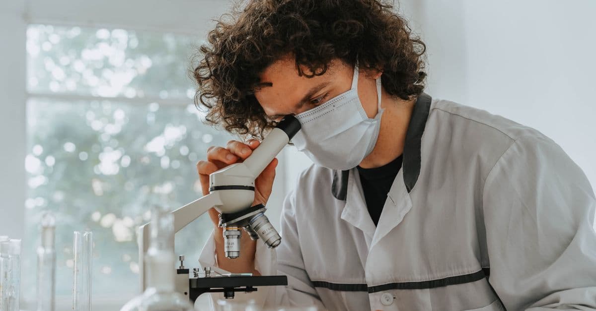 A scientist wearing a face mask examines samples under a microscope in a laboratory setting.