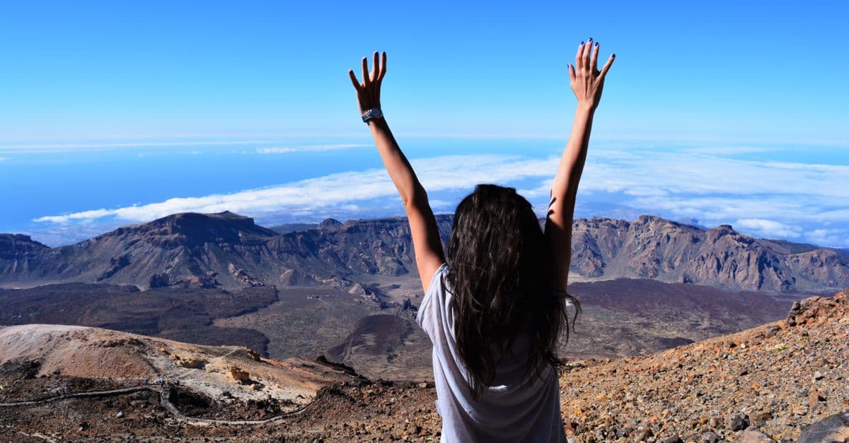 A woman celebrates atop Mount Teide with arms raised, overlooking the vast landscape and sea.