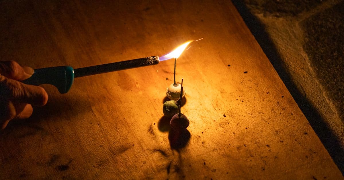 Close-up of a hand lighting a stack of firecrackers on a wooden surface using a kitchen lighter in low light.