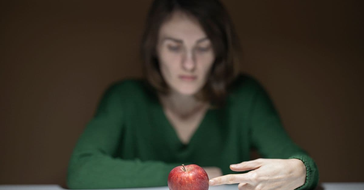 A woman in green thoughtfully observes a red apple on a white table indoors.
