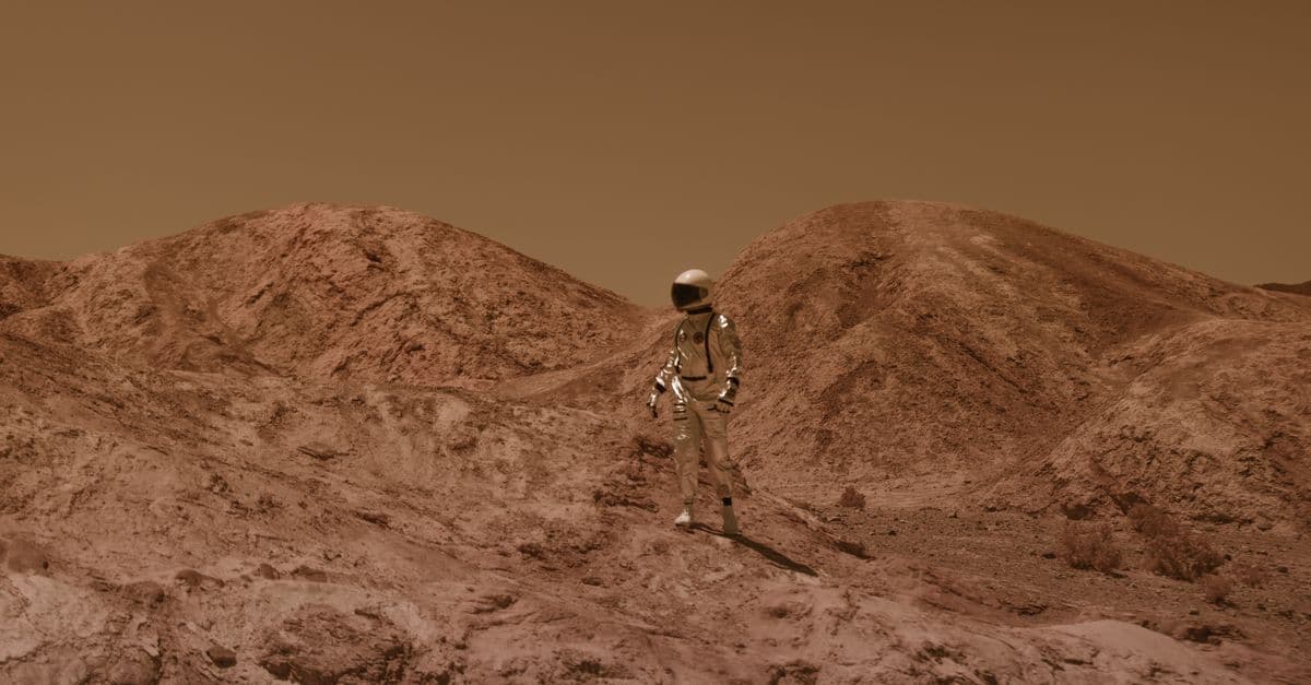 Astronaut in spacesuit explores barren, Mars-like desert terrain under brown sky.