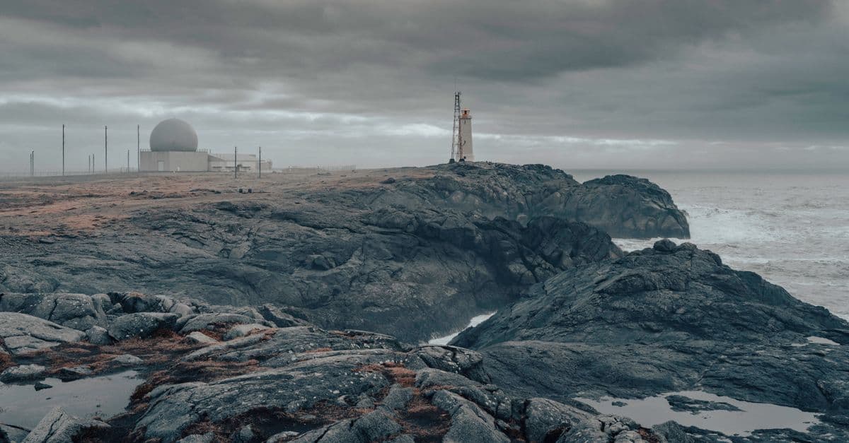 Moody seascape of Iceland's rugged coastline with a lighthouse and a radar dome under cloudy skies.