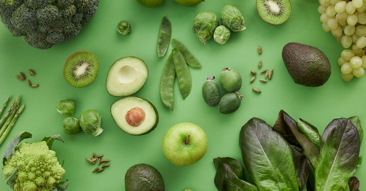 Top view of assorted fresh green vegetables and fruits on a green background, perfect for healthy lifestyle visuals.