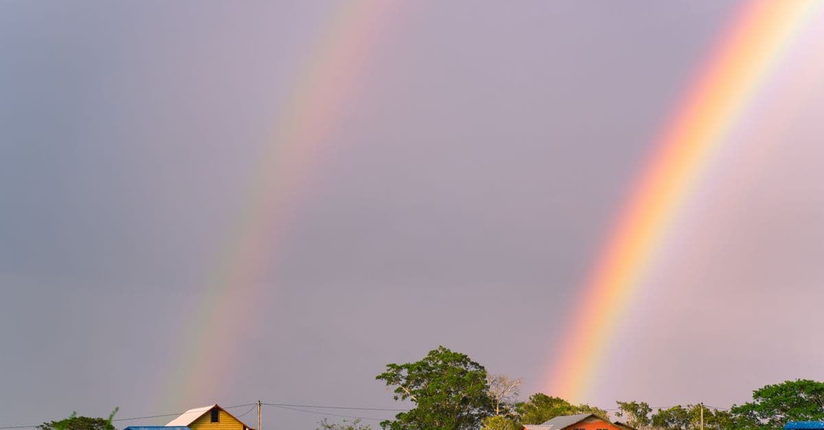 Scenic view of two rainbows arching over rural homes and lush countryside under a cloudy sky.