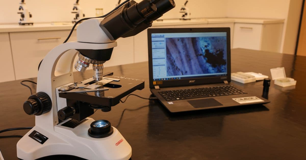 Close-up of a microscope connected to a laptop in a laboratory environment.
