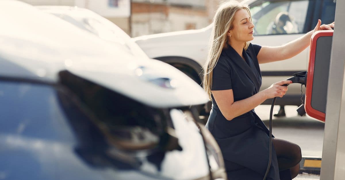 Side view of full length female driver in casual dress squatting near station while charging electric automobile for continuation of driving