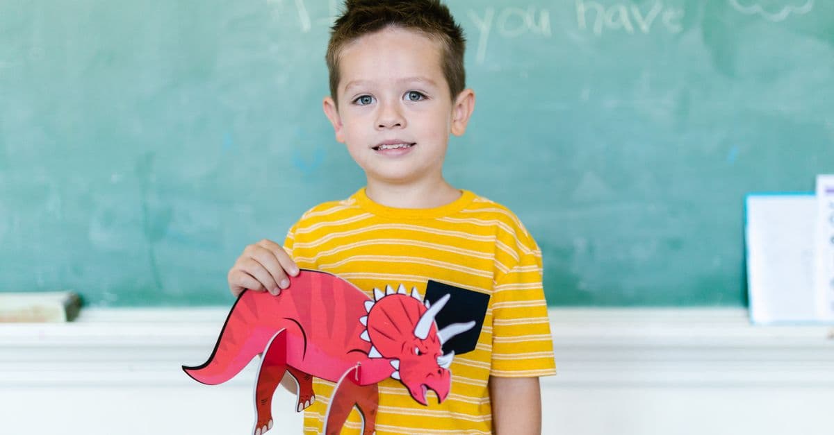 A young boy proudly displays a red triceratops paper cutout in a classroom setting.