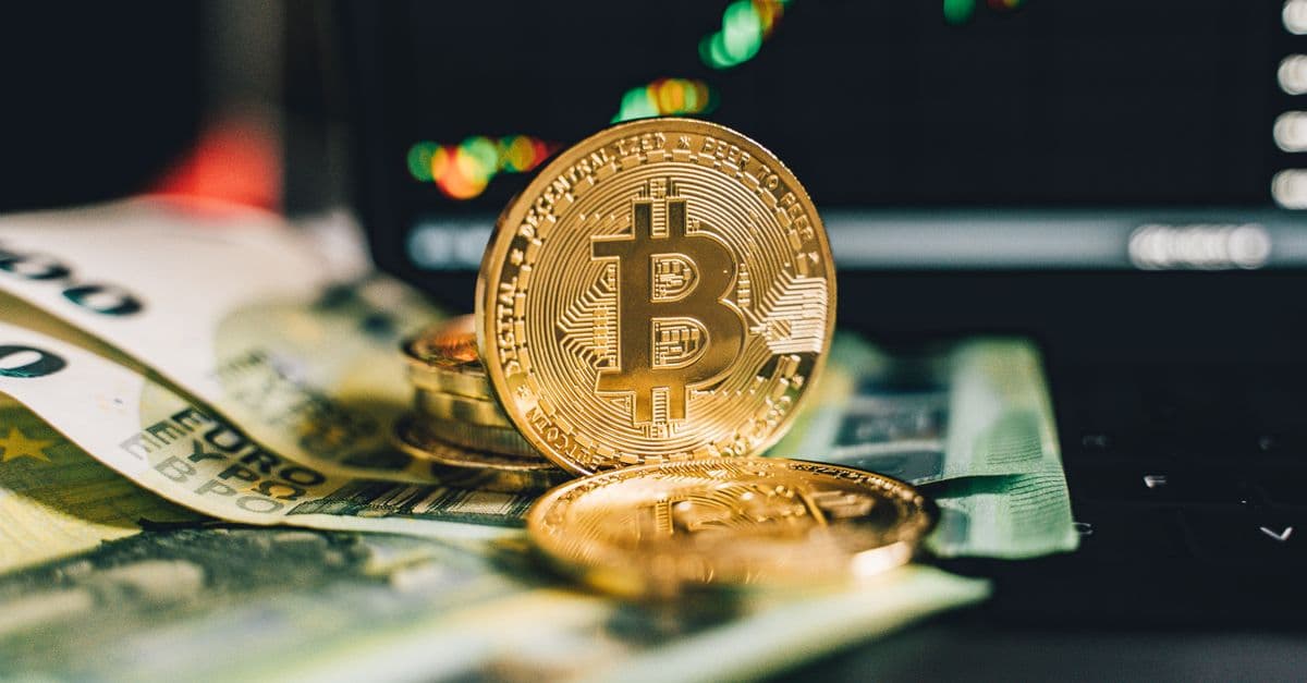 Close-up of Bitcoin and Euro notes on laptop keyboard with market chart in background.