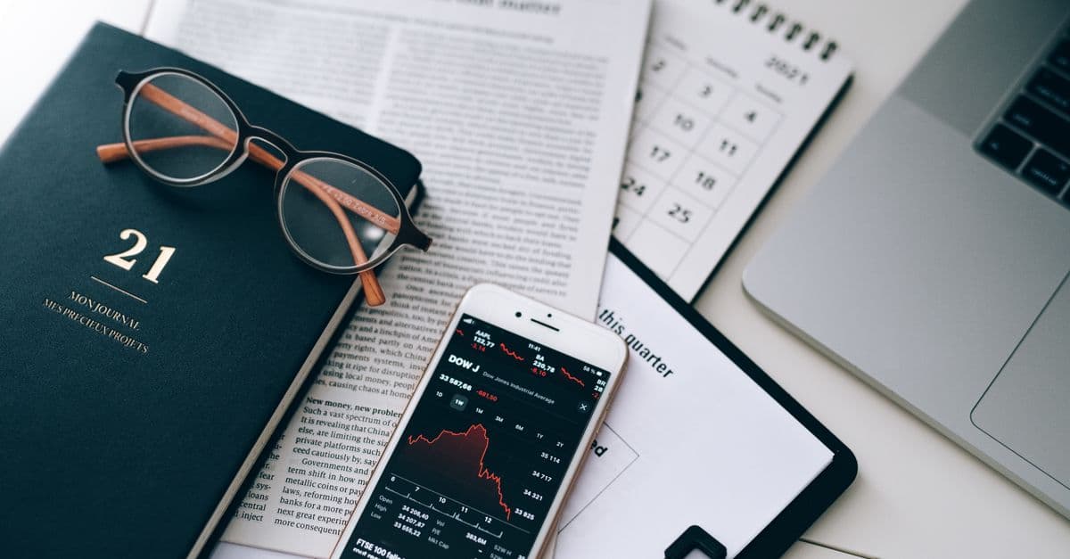 A stylish workspace featuring financial documents, eyeglasses, an iPhone displaying stock data, and a laptop.