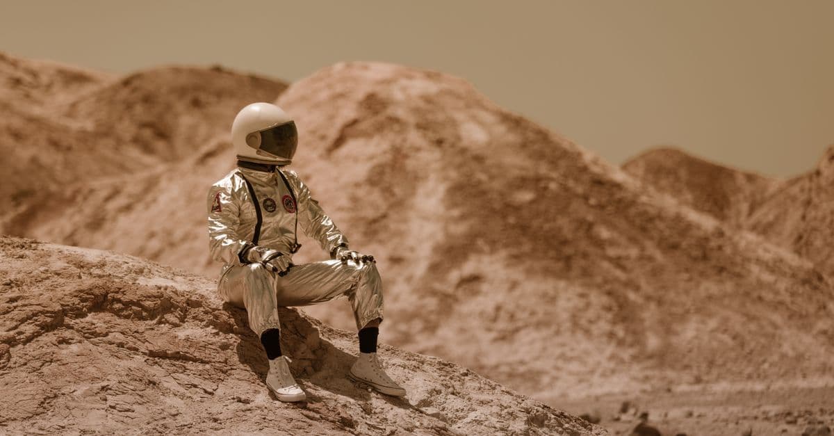 An astronaut in full space suit sits on a rocky desert terrain under a clear sky.