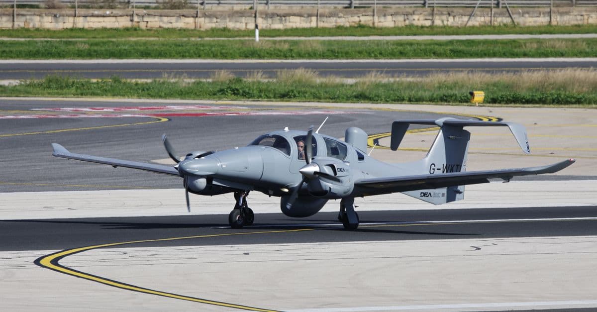 Twin-engine aircraft taxiing on airport runway with pilot visible inside cockpit.