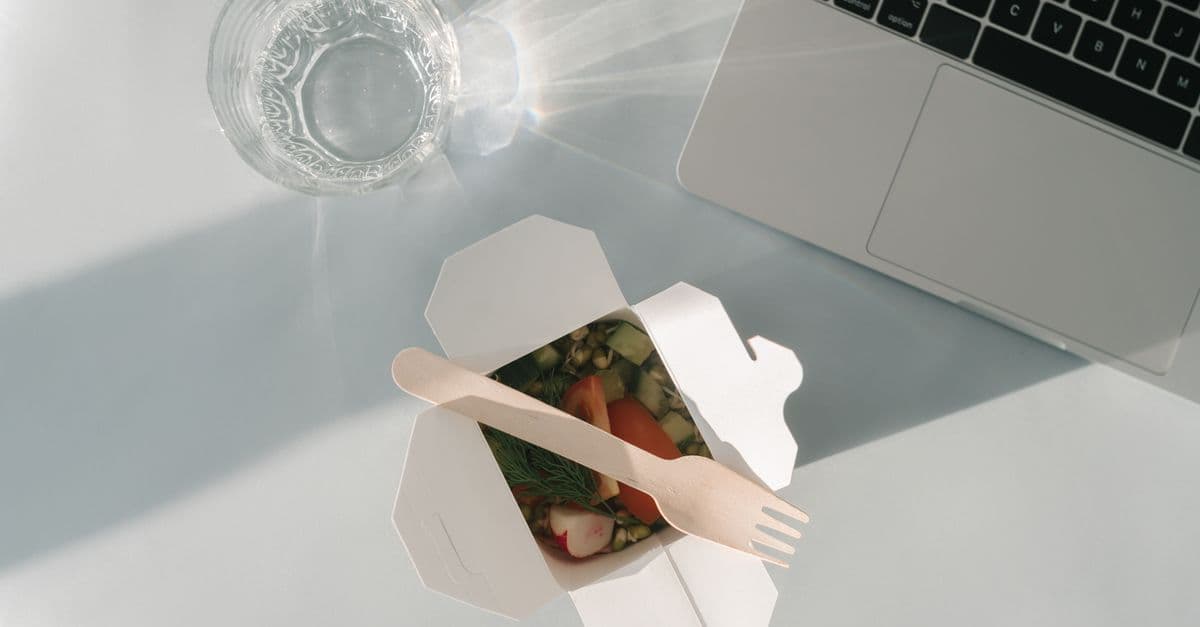 Healthy salad in a takeout box with a glass of water and a laptop on a white surface, top view.