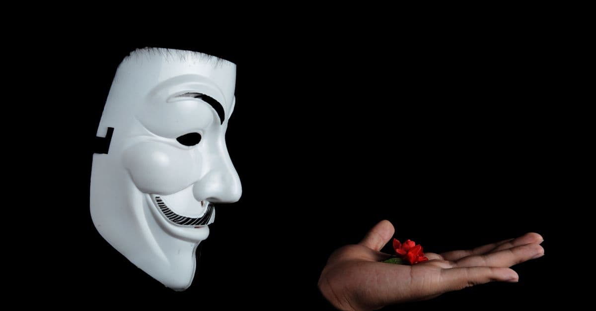 Side view of an anonymous mask facing a hand holding a red flower against a black background.