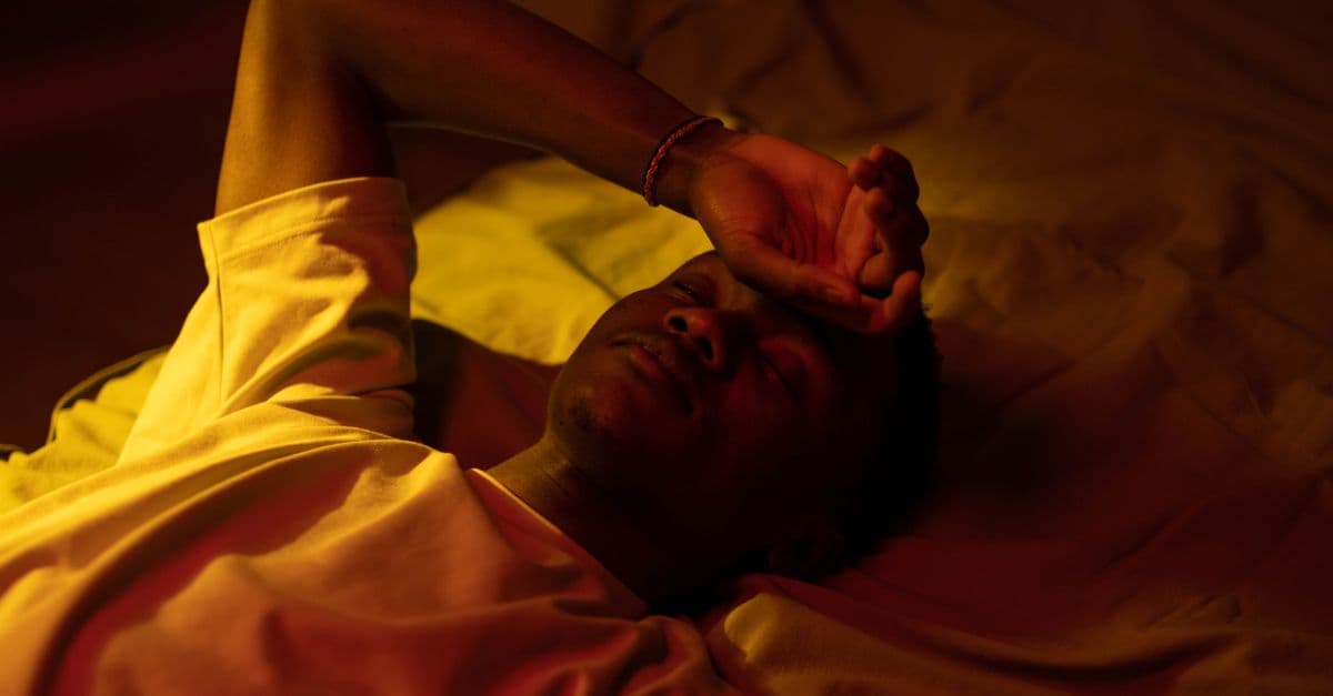 A man lying in bed at night with a moody expression and warm lighting.