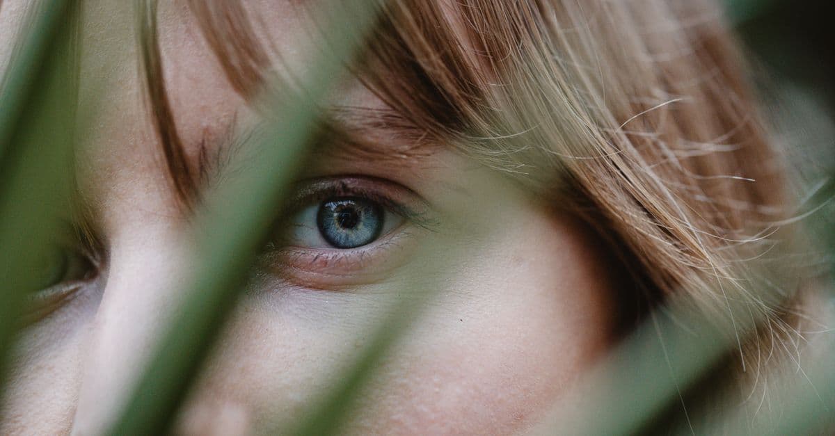A close-up shot of a blue eye partially hidden behind green leaves.