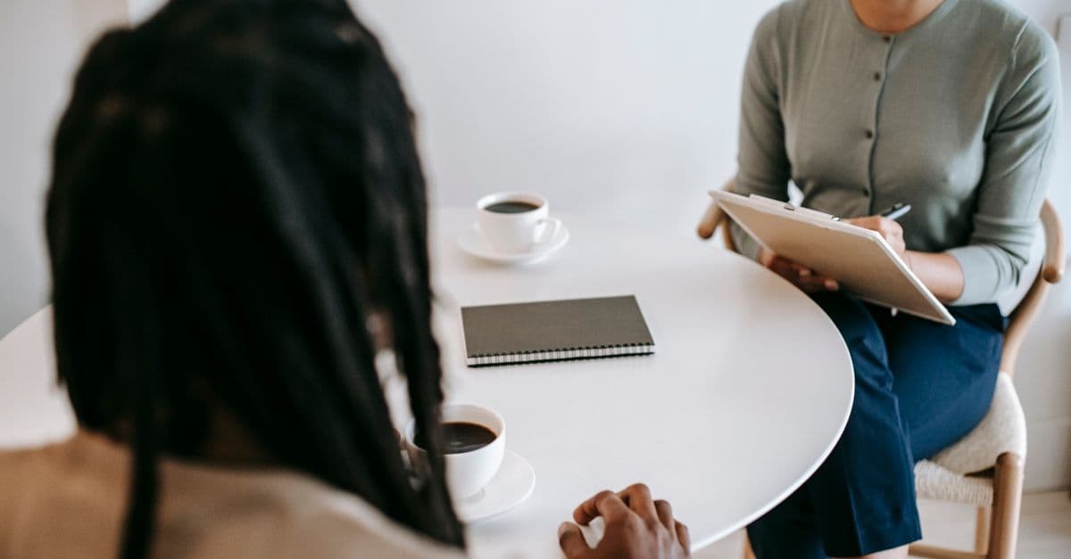 Professional ethnic female psychotherapist asking questions to black male patient and taking notes in clipboard while sitting together at table in modern psychotherapy center