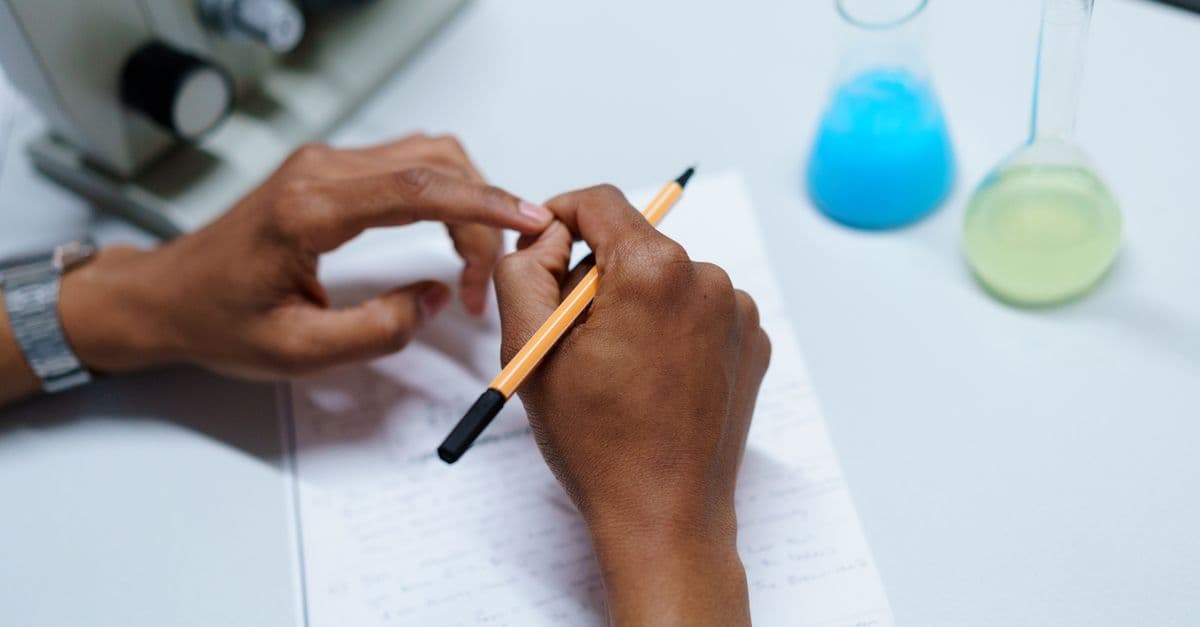 Close-up of a scientist taking notes in a laboratory setting with flasks and a microscope.