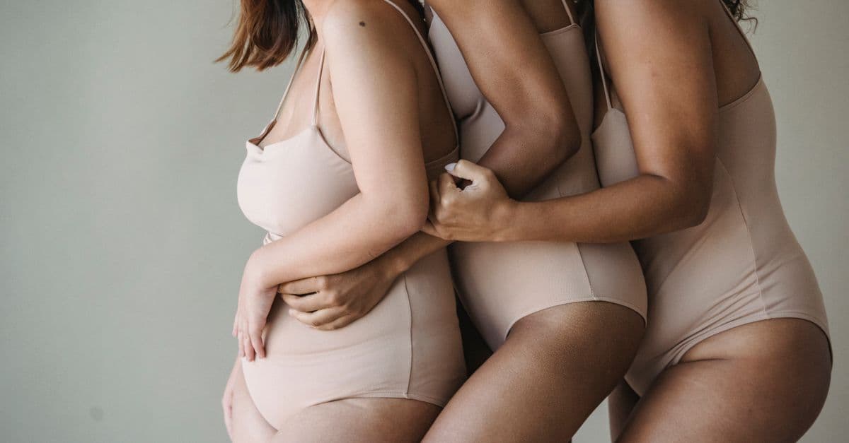 Three diverse women embracing, promoting body positivity and diversity in neutral lingerie.