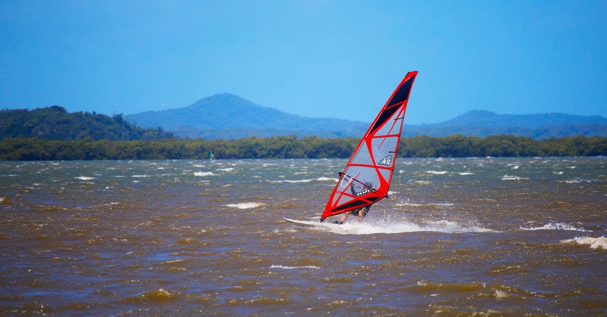 A windsurfer navigates the waves with a vivid sail against a scenic mountain backdrop.
